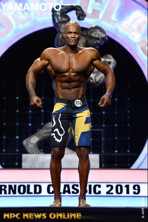 Antony Bessala in a front pose on the Arnold Classic 2019 stage, wearing blue and yellow shorts