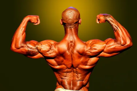 Antony Bessala seen from behind in a back double biceps, olive-gold studio background