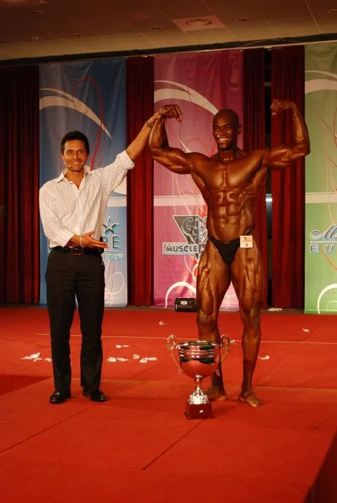 Antony Bessala with his arm raised on stage next to a large trophy under a Musclemania Europe banner