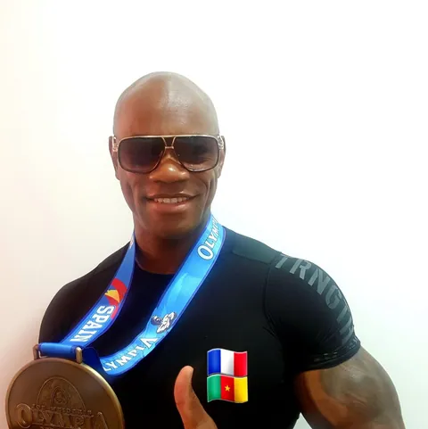 Antony Bessala in sunglasses showing an Olympia Spain medal, with France and Cameroon flag pins on his shirt