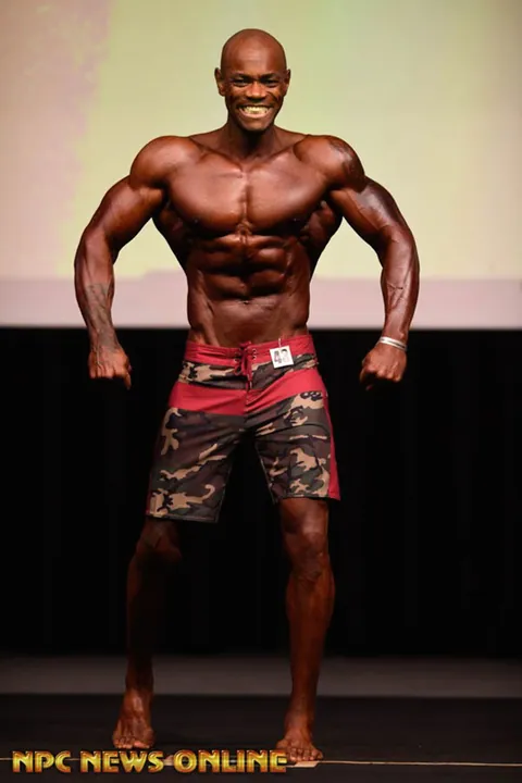 Antony Bessala on a theatre stage in red camouflage shorts, front pose