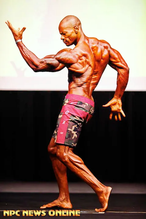 Antony Bessala in a graceful side pose on stage in red camouflage shorts
