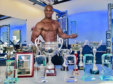 Antony Bessala behind a table of trophies including a Musclemania plaque from Las Vegas
